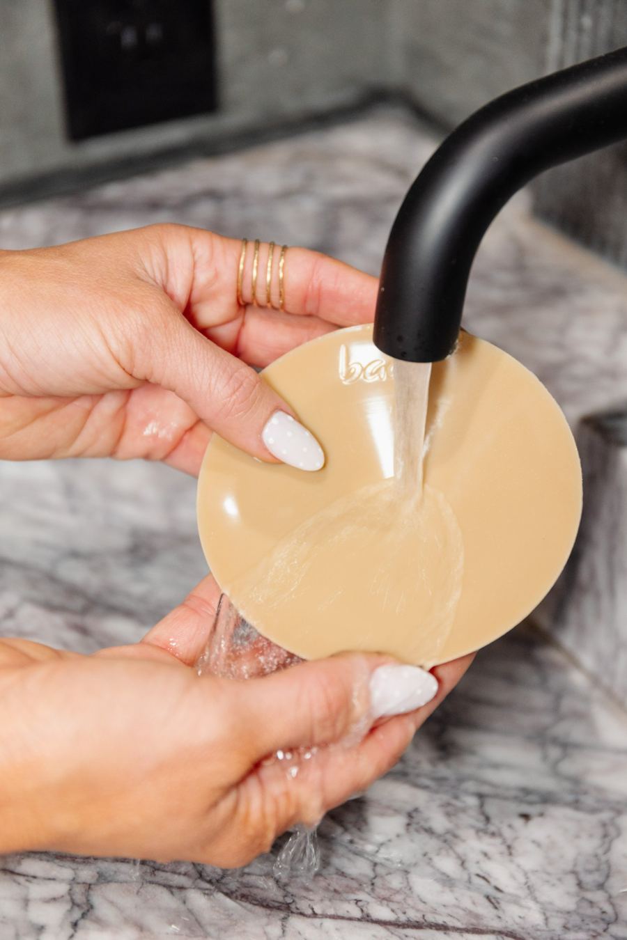Hands rinsing a nude Baae cover under a black faucet over a marble sink